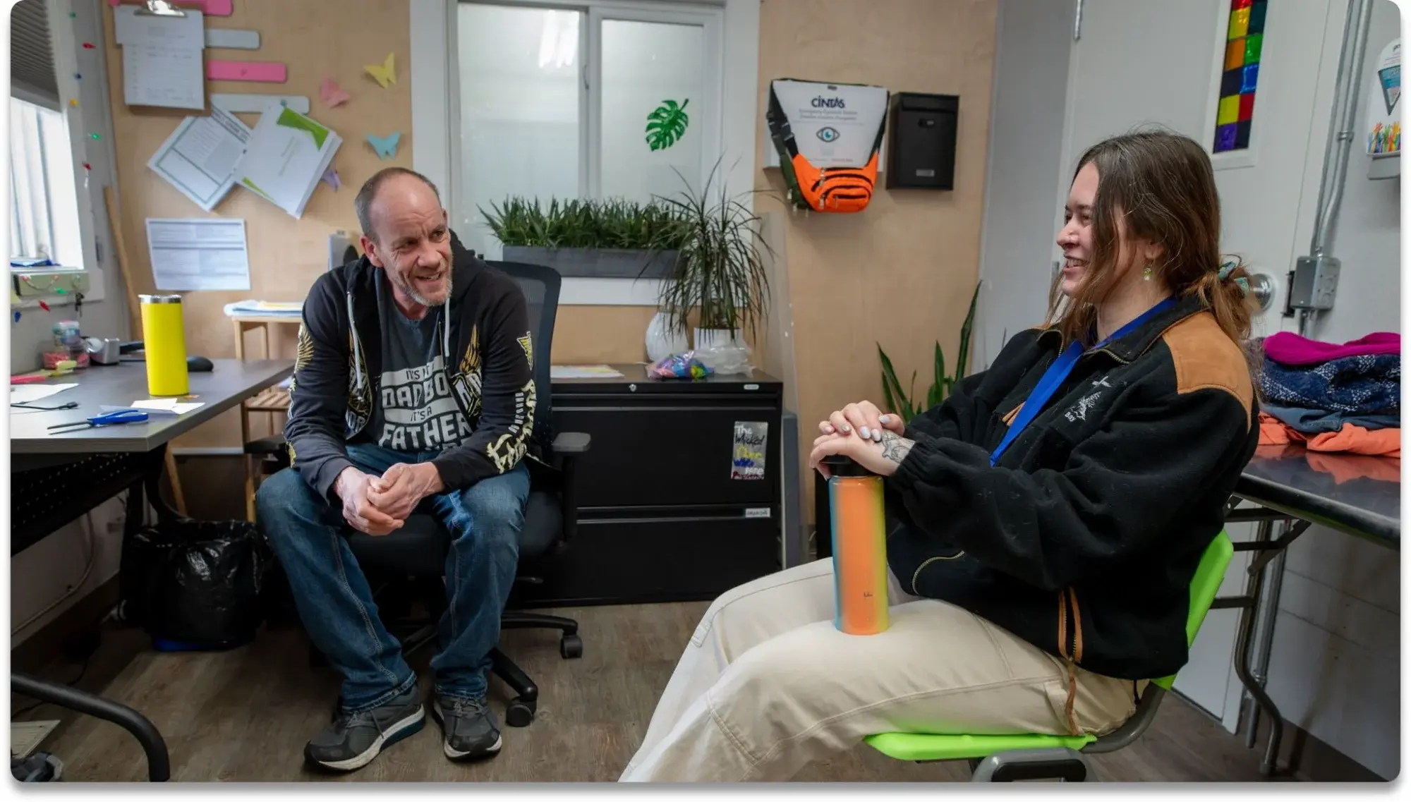 Two people having a friendly conversation in a community office space; an older man with a goatee wearing a graphic t-shirt sits on an office chair while a young woman with long brown hair in a black and tan jacket sits across from him holding a colorful water bottle, surrounded by plants, bulletin boards with papers and origami decorations.
