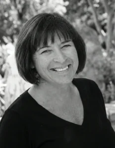 Black and white portrait of a woman with a short dark bob haircut and bangs, smiling broadly at the camera, wearing a black v-neck top, photographed outdoors with lush garden foliage in the soft-focus background