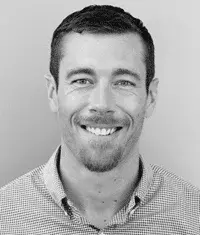 Black and white professional headshot of a man with short hair and a beard, smiling warmly at the camera, wearing a casual collared shirt against a plain gray background