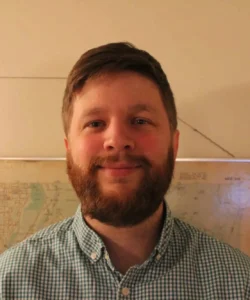 Man with short brown hair and a full beard smiling at the camera, wearing a green and white gingham check button-up shirt, standing in front of a framed map of Vancouver on a cream-colored wall.