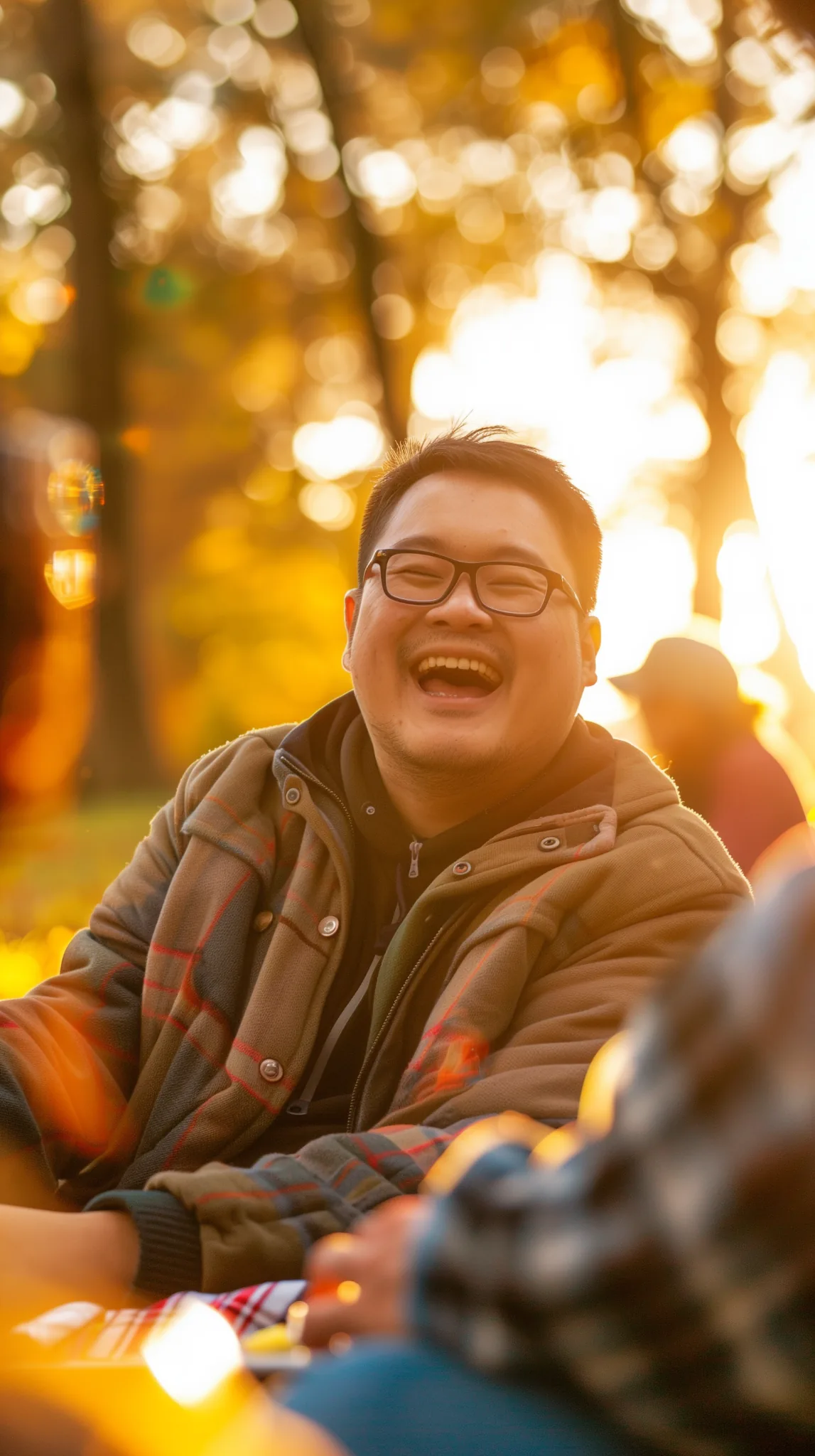 Smiling participant wearing glasses laughs during a community inclusion and outreach gathering in the Okanagan, seated outdoors in warm golden-hour light with others nearby.
