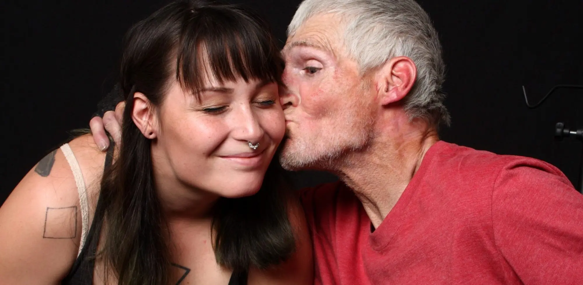 father_daughter_ Tender portrait of an older man with gray hair and beard wearing a red v-neck shirt kissing a younger woman on the cheek, as she smiles with her eyes closed; she has long dark hair with bangs, a septum piercing, and tattoos on her arm, photographed against a dark background.