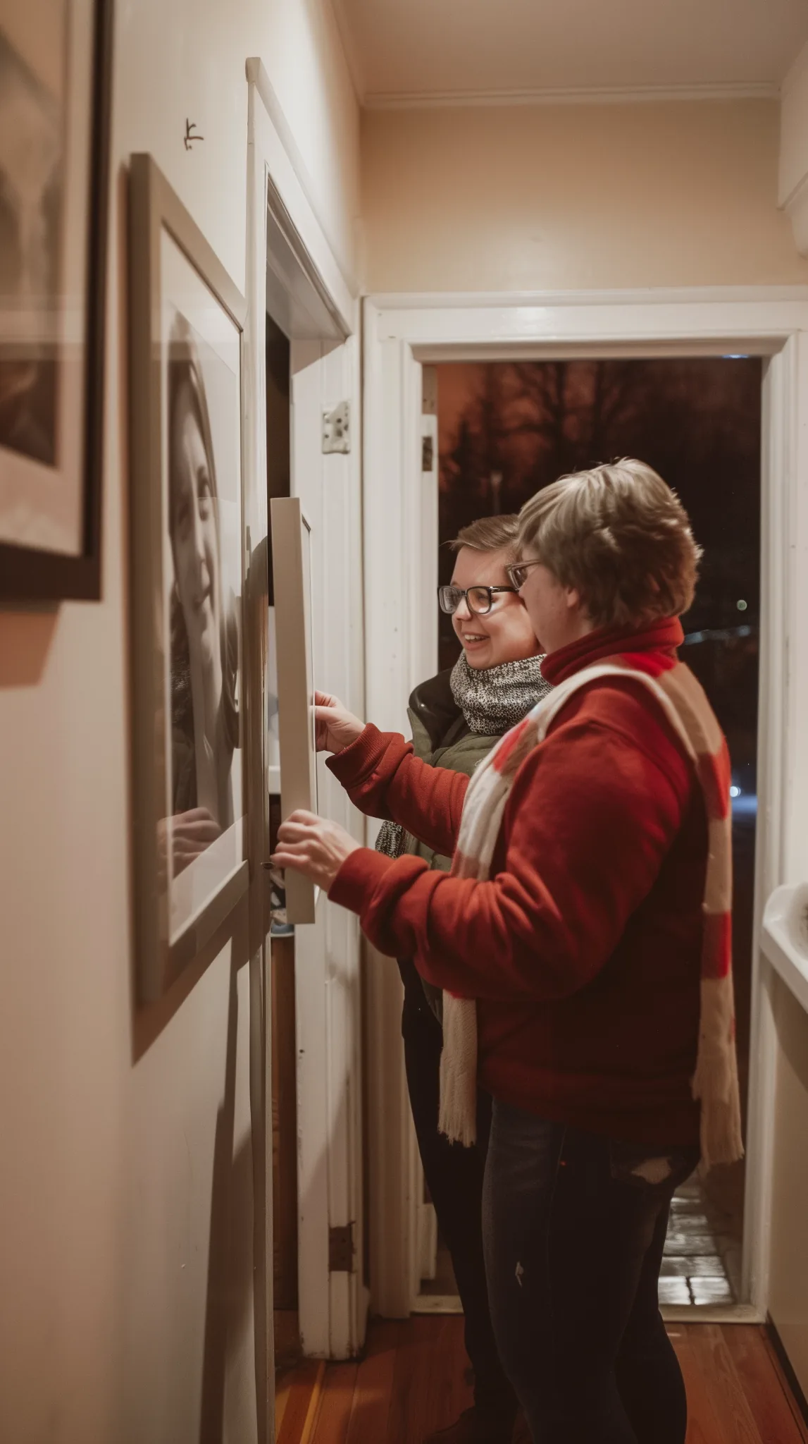 Two people smiling together in a shared home hallway, adjusting framed photos during a Homeshare program visit in the Okanagan, highlighting connection and supportive housing.