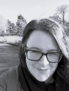 Black and white portrait of a woman with long brown hair and glasses, smirking at the camera, taken outdoors with trees and grass in the background.