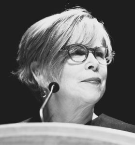 Black and white portrait of a woman with a short grey bob haircut and glasses, looking to the left, standing behind a podium with a microphone.